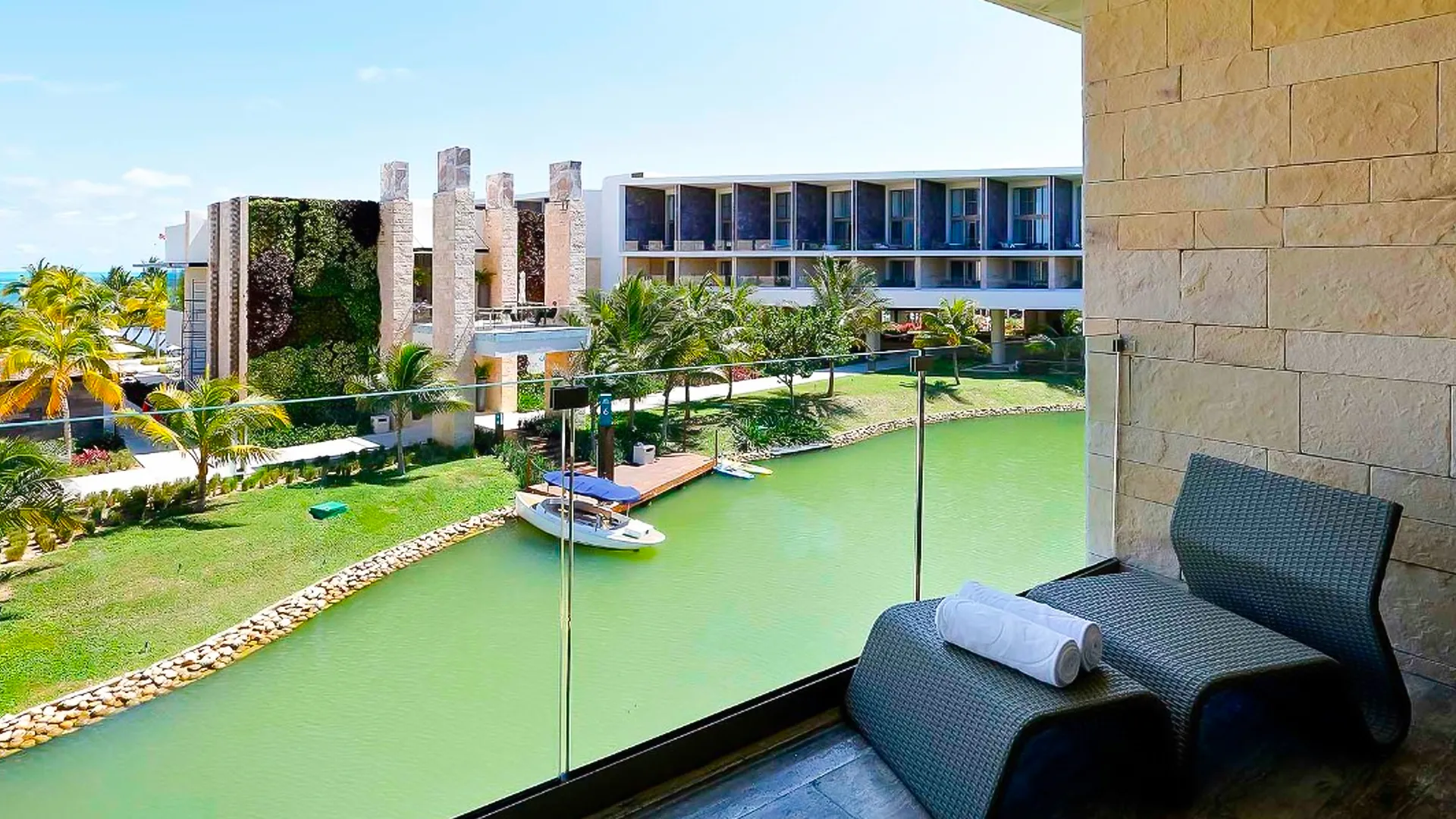 A private balcony at Grand Palladium Cancun Resort with a cushioned gray lounge chair, a view of a lagoon, tropical greenery, and resort buildings in the background.