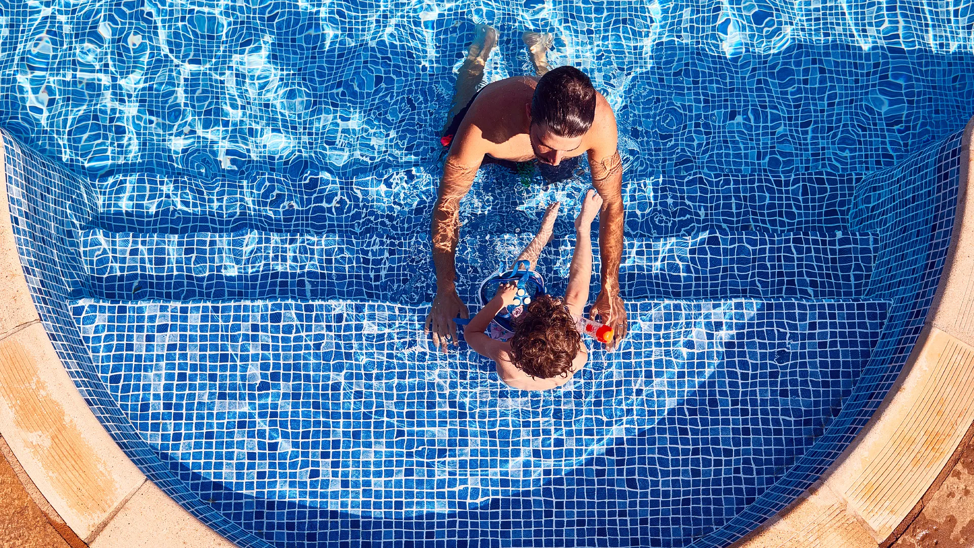 A person in a pool assisting a child in the water, with both individuals partially submerged, surrounded by blue tiled pool edges under bright daylight.