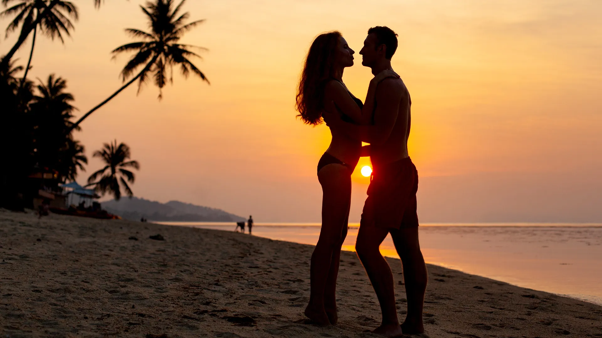 A silhouette of a couple embracing on a beach at Hyatt Ziva Cancun during sunset, with palm trees and the sun low on the horizon.