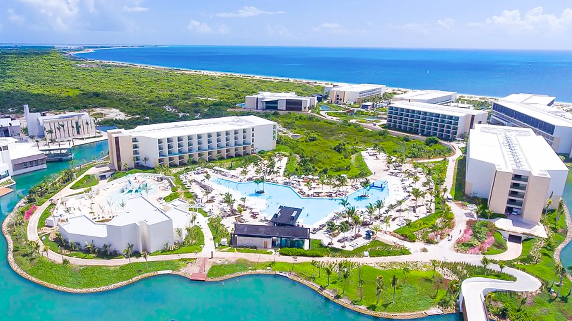 An aerial view of Grand Palladium Cancun Resort featuring multiple white multi-story buildings, a large central pool, lush tropical greenery, and the ocean in the background.