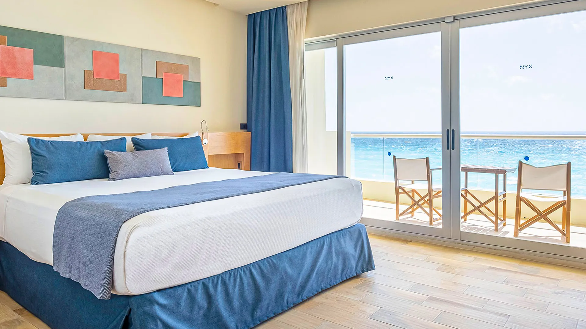 Luxurious hotel room with a king-size bed, blue bedding, and abstract wall art, featuring sliding glass doors to a balcony with chairs overlooking the Caribbean Sea, wooden floors and modern decor.