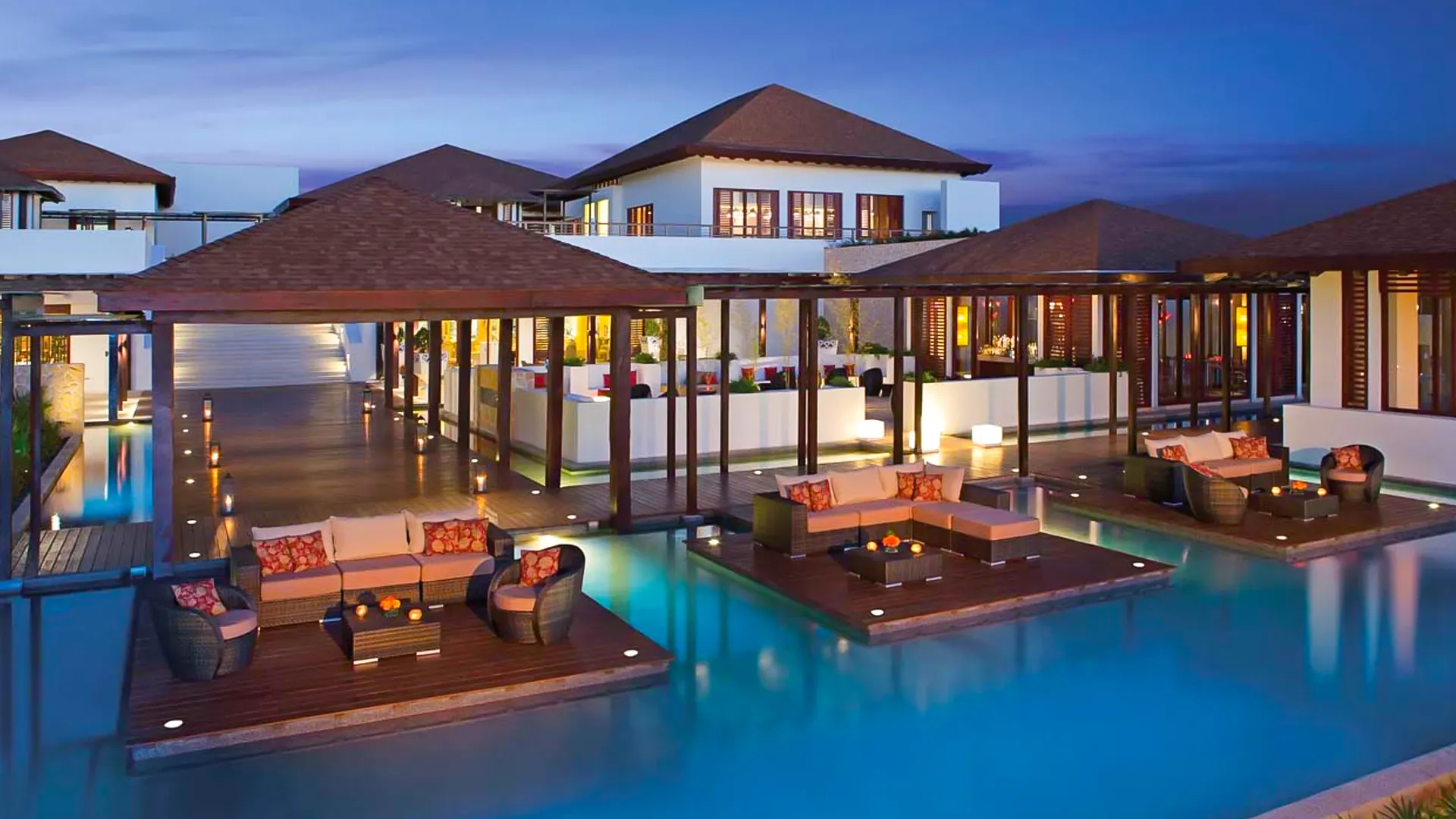 A luxurious evening scene of a resort with floating wooden platforms over a pool, featuring cozy seating areas with colorful cushions and ambient lighting.