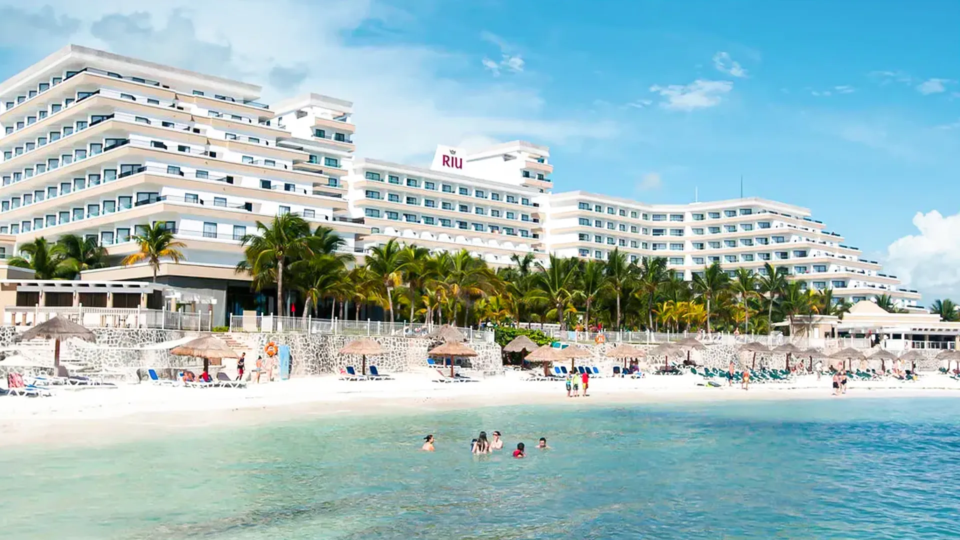Beachfront view of Riu Caribe Cancun with palm trees, sun loungers, and guests swimming in clear turquoise water.
