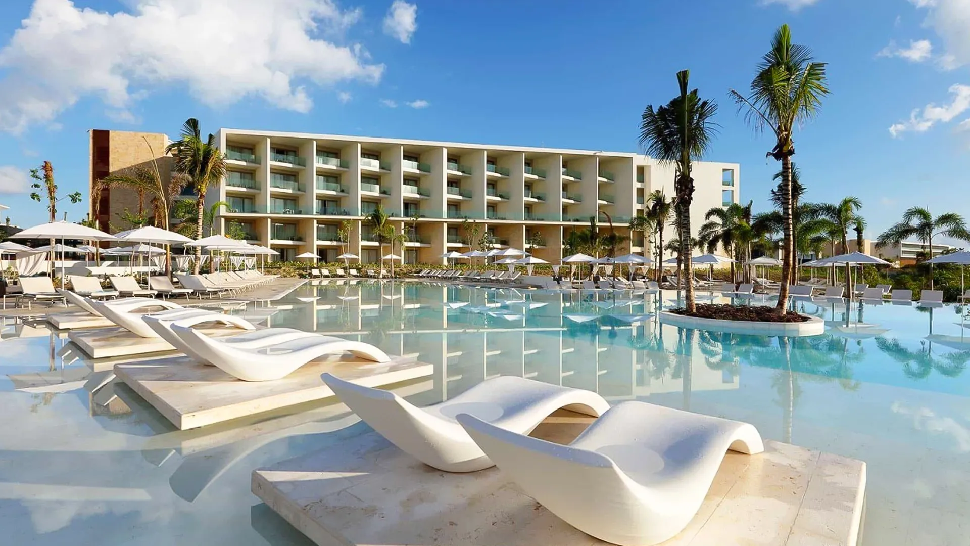 A luxurious pool area at Grand Palladium Cancun Resort featuring multiple white lounge chairs on platforms in shallow water, with a modern multi-story building, palm trees, and a clear blue sky in the background.