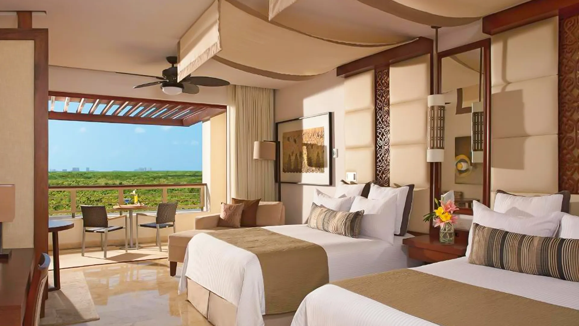 Spacious resort bedroom featuring two queen beds with neutral linens, wooden accents, a ceiling fan, sitting area, and large windows opening to a balcony with views of lush green fields and blue sky.