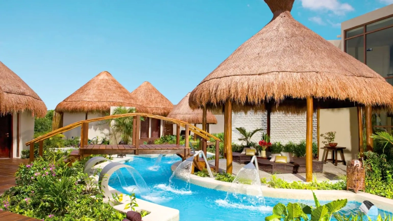 Serene tropical pathway at Dreams Riviera Cancun resort, featuring multiple thatched-roof palapa bungalows connected by a wooden arched bridge over a winding blue water channel with cascading fountains, lush green foliage, potted plants, and lounge seating under a clear blue sky.