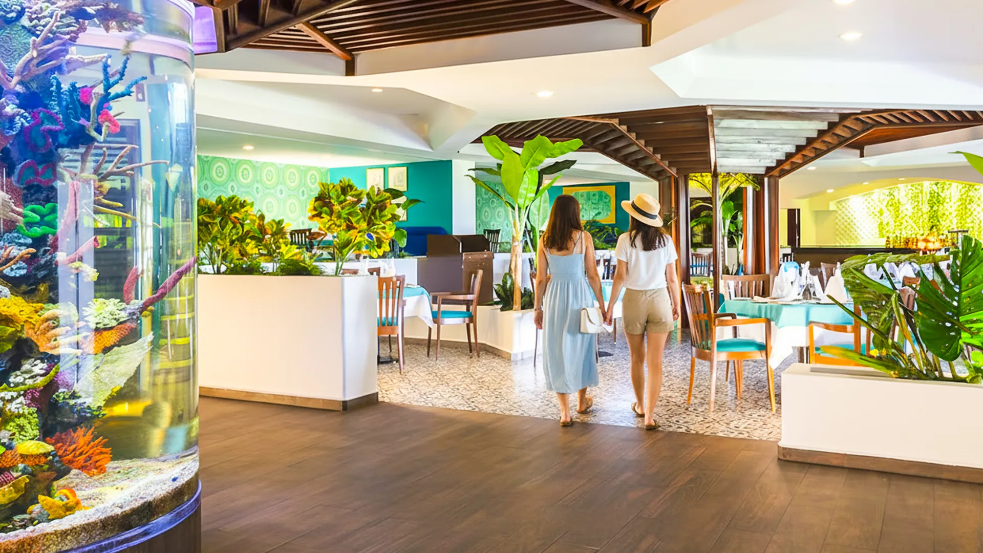 Two women in light blue dresses standing in a modern indoor lounge at Crown Paradise Club Cancun, with a cylindrical aquarium filled with colorful fish, white furniture, potted plants, and a bright open space with natural light.