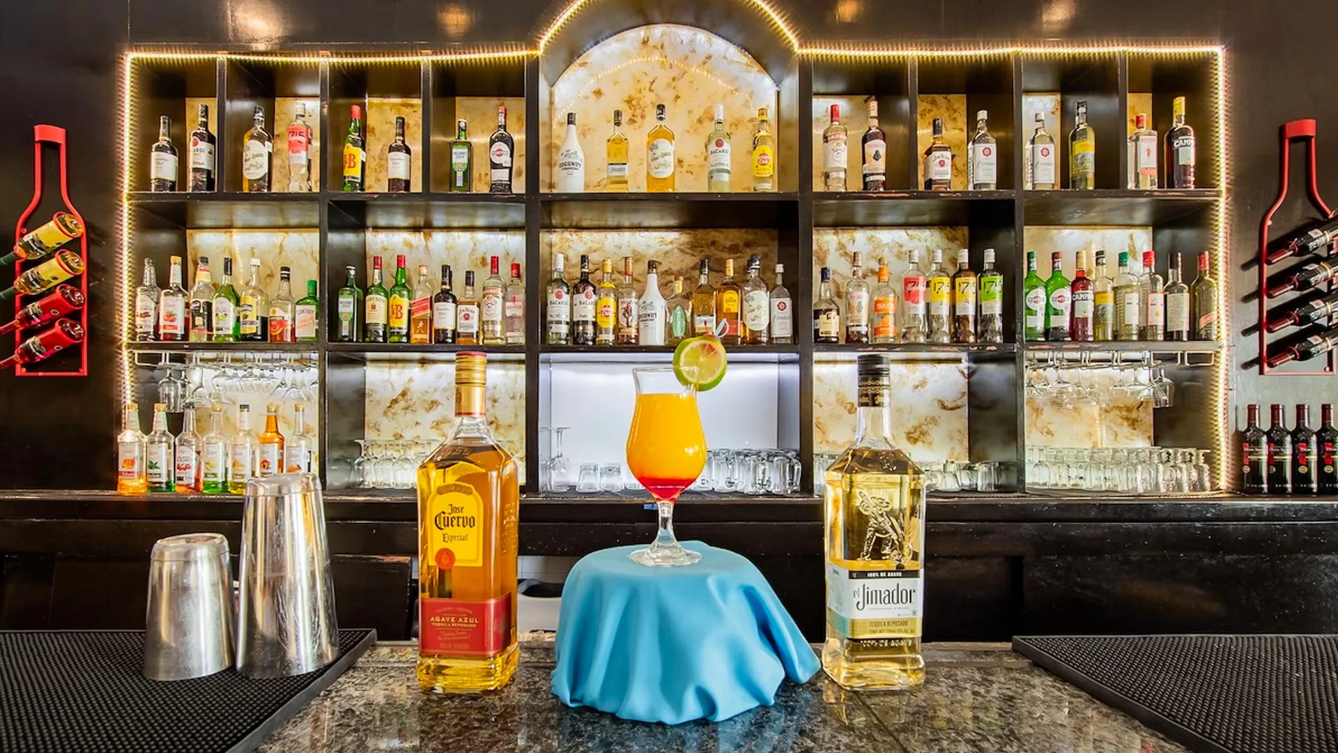 Bar area at the GR Solaris Cancun resort with a well-stocked shelf of liquor bottles, a cocktail with lime garnish, and two tequila bottles prominently displayed on the counter.