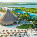 Aerial view of Club Med Cancun resort in Riviera Maya, showcasing a large thatched-roof beach club structure on white sand, multiple lounge chairs with umbrellas, palm trees, tennis courts, a lagoon with sailboats, and surrounding mangroves and turquoise waters under a clear blue sky.