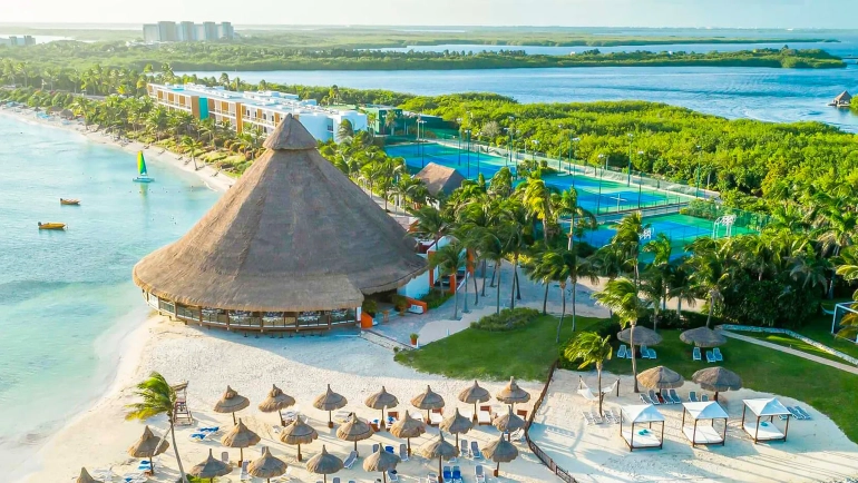 Aerial view of Club Med Cancun resort in Riviera Maya, showcasing a large thatched-roof beach club structure on white sand, multiple lounge chairs with umbrellas, palm trees, tennis courts, a lagoon with sailboats, and surrounding mangroves and turquoise waters under a clear blue sky.
