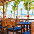 Open-air beachfront restaurant with wooden tables and blue-cushioned chairs under a thatched roof, surrounded by palm trees, red hibiscus flowers, and the turquoise Caribbean Sea in the background.