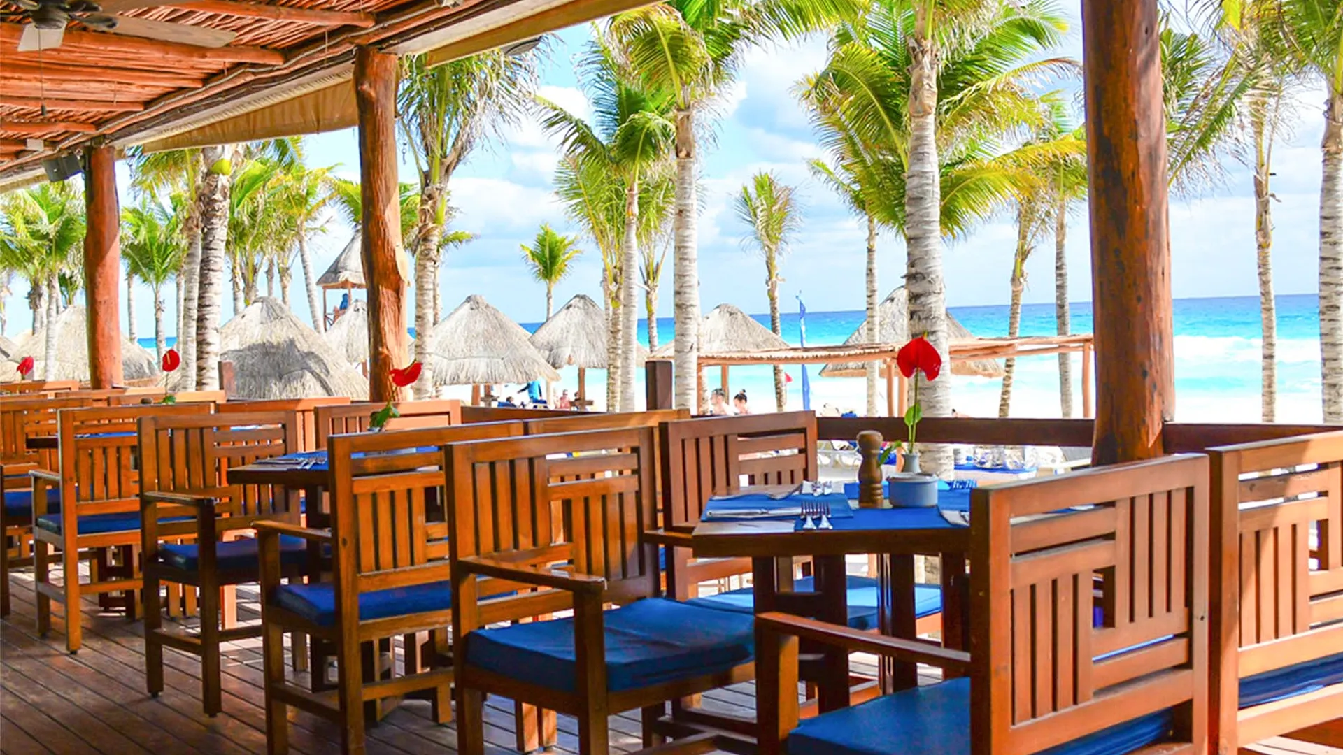 Open-air beachfront restaurant with wooden tables and blue-cushioned chairs under a thatched roof, surrounded by palm trees, red hibiscus flowers, and the turquoise Caribbean Sea in the background.