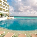 A luxurious infinity pool at a resort with clear blue water, surrounded by lounge chairs and a multi-story building with balconies, overlooking the turquoise ocean under a partly cloudy sky.