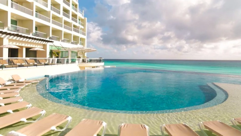 A luxurious infinity pool at a resort with clear blue water, surrounded by lounge chairs and a multi-story building with balconies, overlooking the turquoise ocean under a partly cloudy sky.