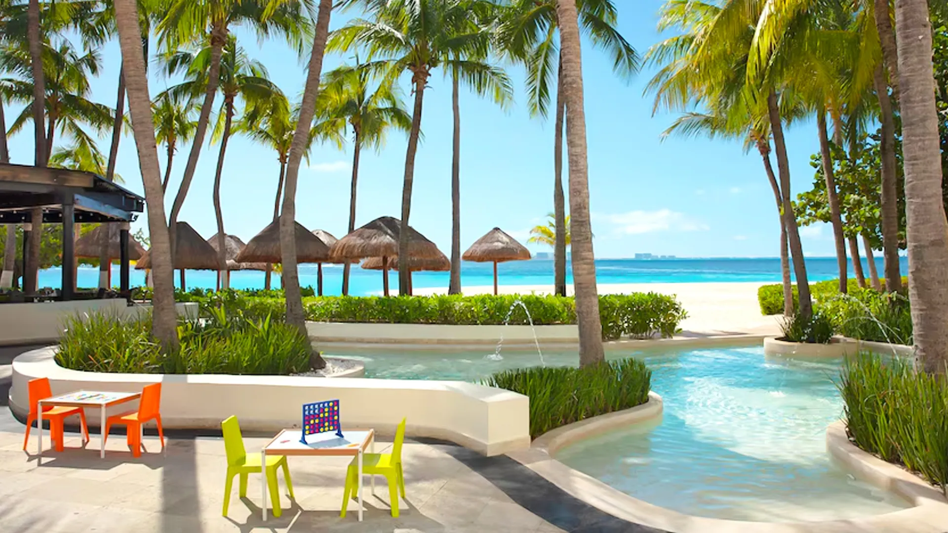 A family-friendly outdoor play area at a Cancun resort, showcasing a curved white infinity pool with clear water, lush green landscaping, several thatched-roof cabanas, orange and green plastic chairs around small tables with a Connect Four game, tall palm trees, and a white sandy beach with turquoise ocean in the background under a sunny sky.