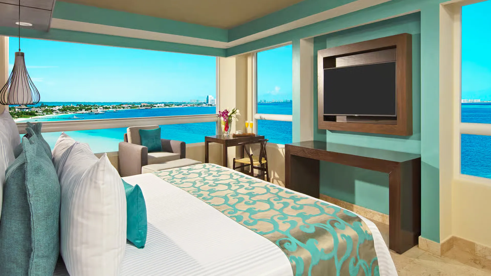 A luxurious oceanfront hotel room in a Cancun resort, featuring a king-size bed with teal and white bedding, teal accent walls, a flat-screen TV mounted on wood paneling, a small side table with champagne flutes and flowers, a gray armchair, and expansive floor-to-ceiling windows offering panoramic views of the turquoise sea and distant city skyline under a bright blue sky.
