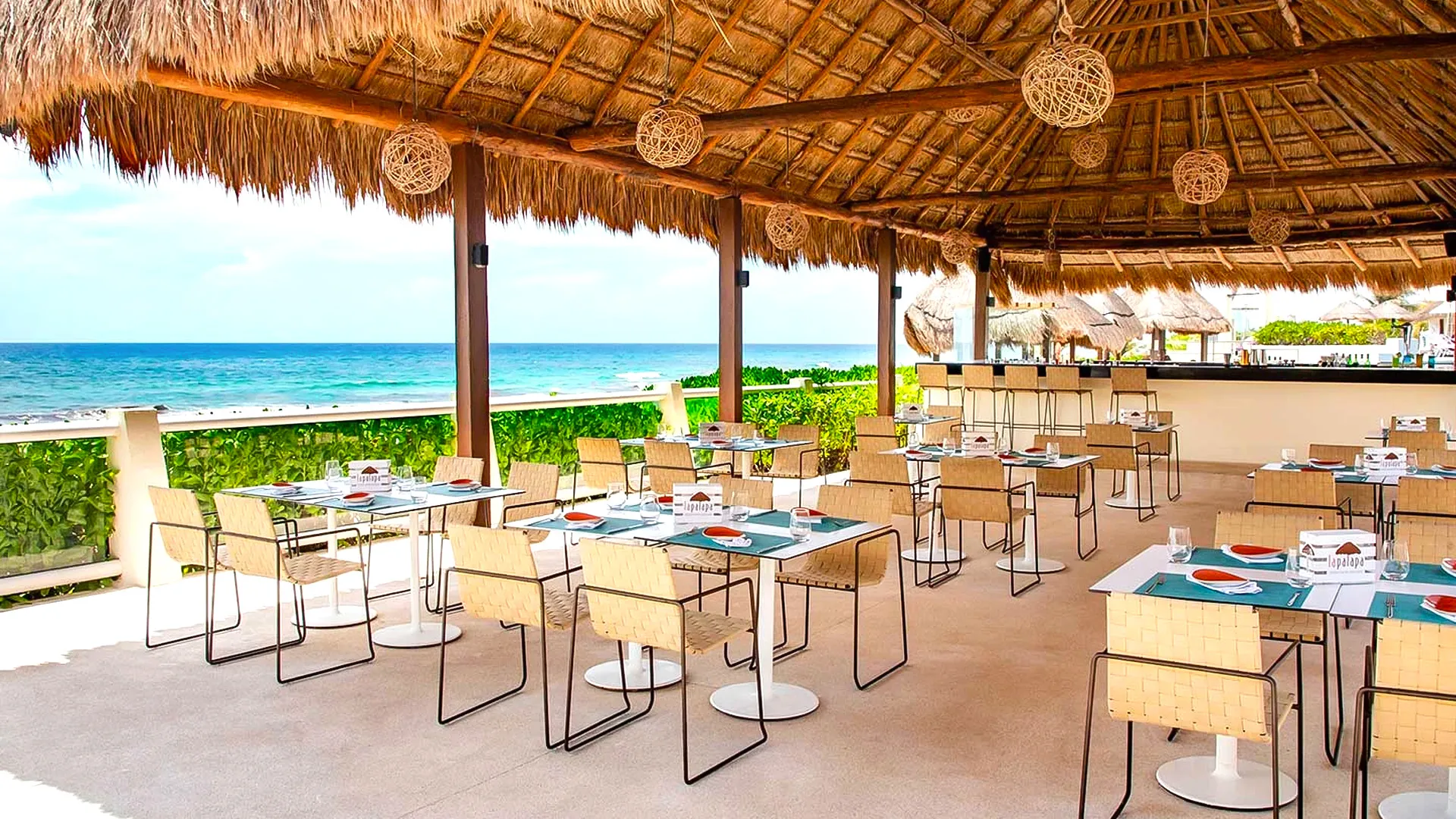 Open-air beachfront restaurant at Crown Paradise Club Cancun under a thatched palapa roof, featuring wooden tables with blue placemats, chairs, and a panoramic view of the turquoise sea, white sand, and a partly cloudy sky.