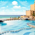 Large infinity-edge pool at The Royal Sands with blue water and white floating noodles, surrounded by beige lounge chairs and thatched umbrellas, palm trees, beige multi-story buildings, and the turquoise Caribbean Sea under a blue sky.