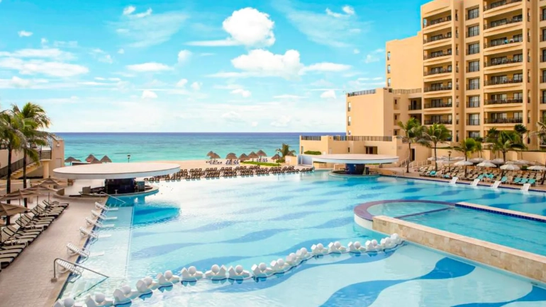 Large infinity-edge pool at The Royal Sands with blue water and white floating noodles, surrounded by beige lounge chairs and thatched umbrellas, palm trees, beige multi-story buildings, and the turquoise Caribbean Sea under a blue sky.