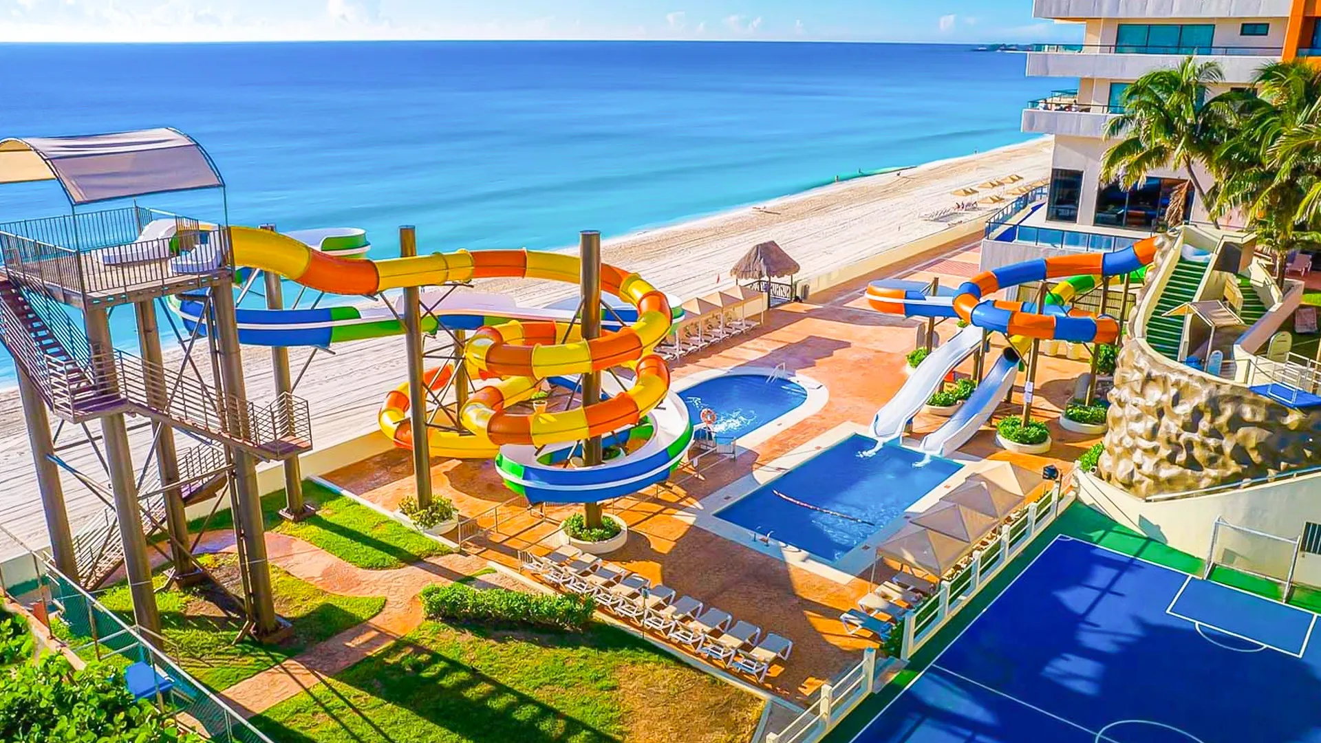 Aerial view of Crown Paradise Club Cancun's colorful water park area, featuring twisting orange and blue spiral slides, a large blue pool, climbing structures, lounge chairs, palm trees, a basketball court, and the white sandy beach and turquoise ocean in the background under a clear blue sky.