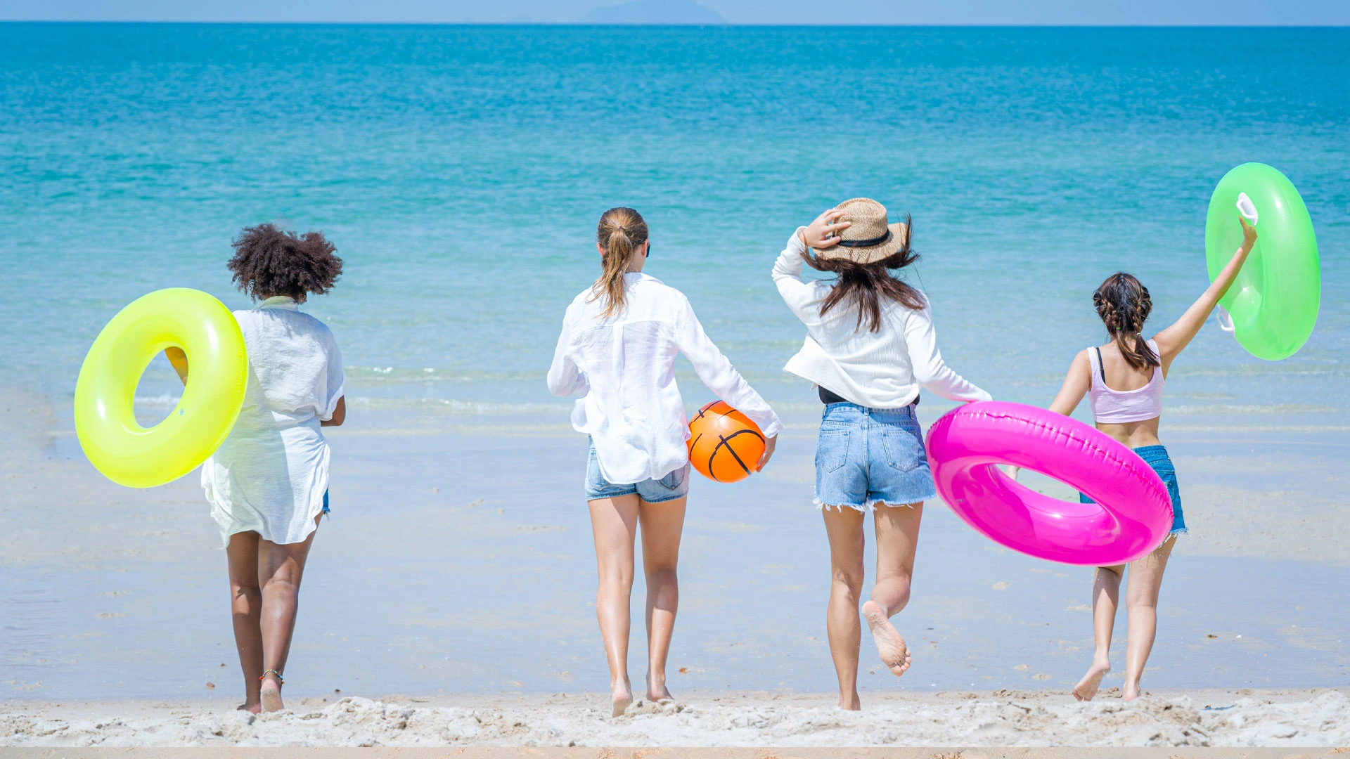 Four young women holding colorful swim rings and a beach ball, walking toward the turquoise ocean at Temptation Cancun Resort.