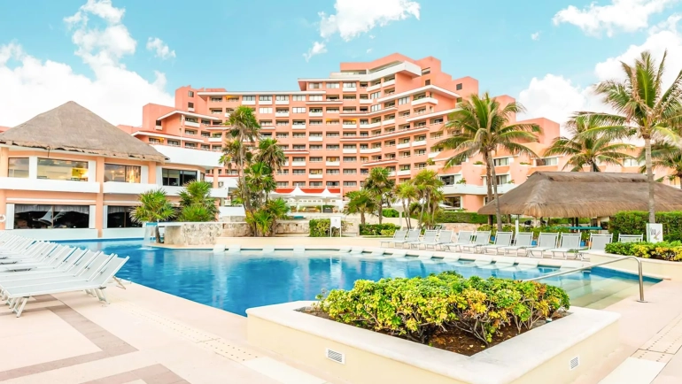 Overview of the Wyndham Cancun resort's pink-hued buildings with thatched-roof structures, a rectangular blue swimming pool lined with white lounge chairs, palm trees, tropical landscaping, and partly cloudy skies.