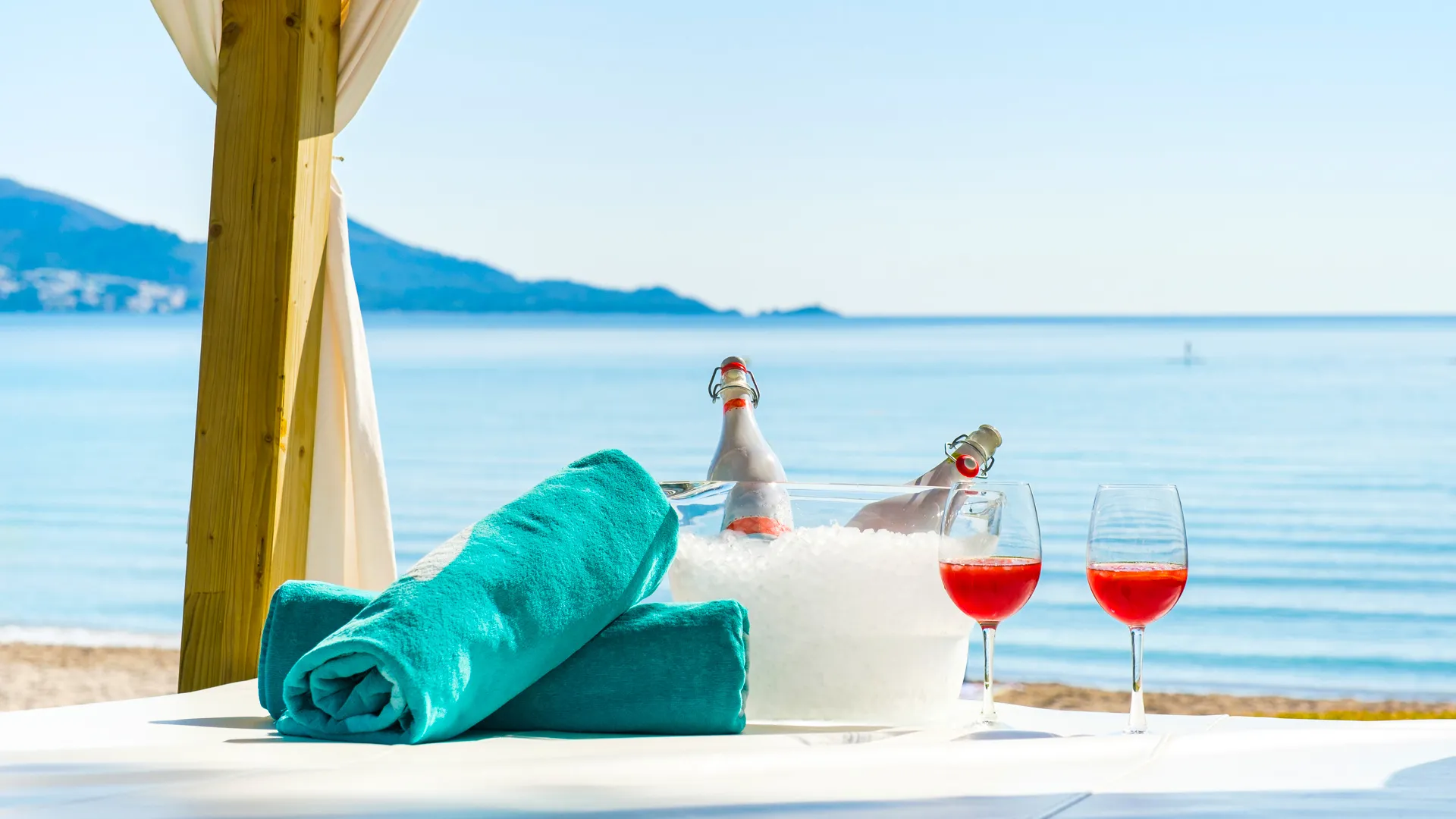 Beach cabana setup with two bottles of rosé champagne in an ice bucket, two glasses of red wine, and stacked turquoise towels on a white table, with calm blue sea and mountains in the distant background.