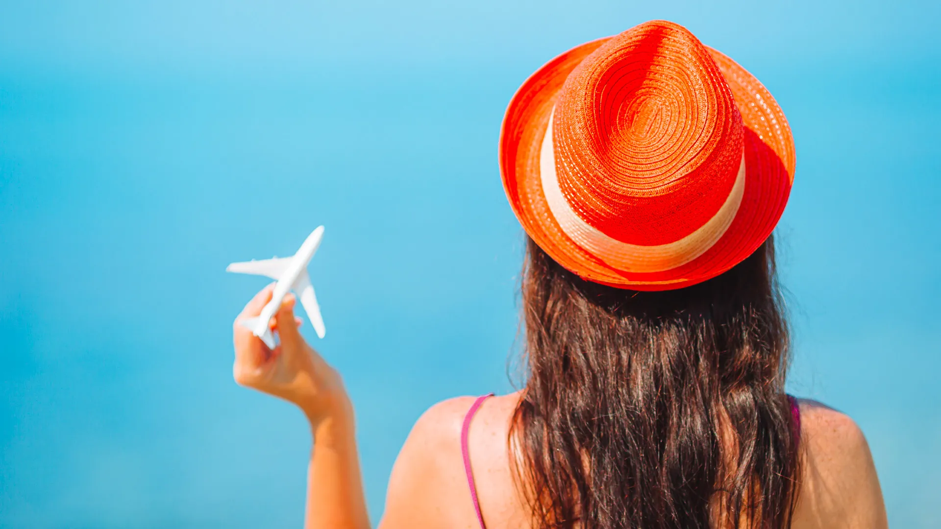 Woman in a red hat holding a small airplane model against a clear blue sky, symbolizing travel dreams.