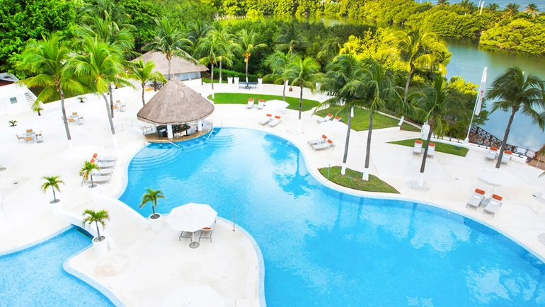 Aerial view of Le Blanc Cancun resort pools winding through lush green landscapes with palm trees, thatched-roof bars and cabanas, white lounge areas, and a serene lagoon under a clear blue sky.
