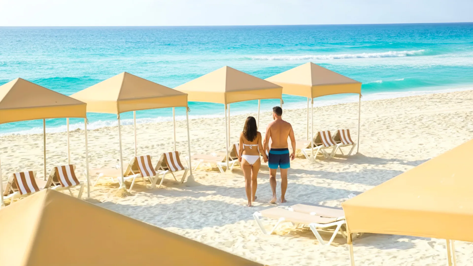 A couple in swimwear—a woman in a black bikini and a man in blue trunks—standing on a white sandy beach at Crown Paradise Club Cancun, with yellow umbrellas and lounge chairs arranged along the shoreline, and the turquoise ocean waves in the background under a clear blue sky.