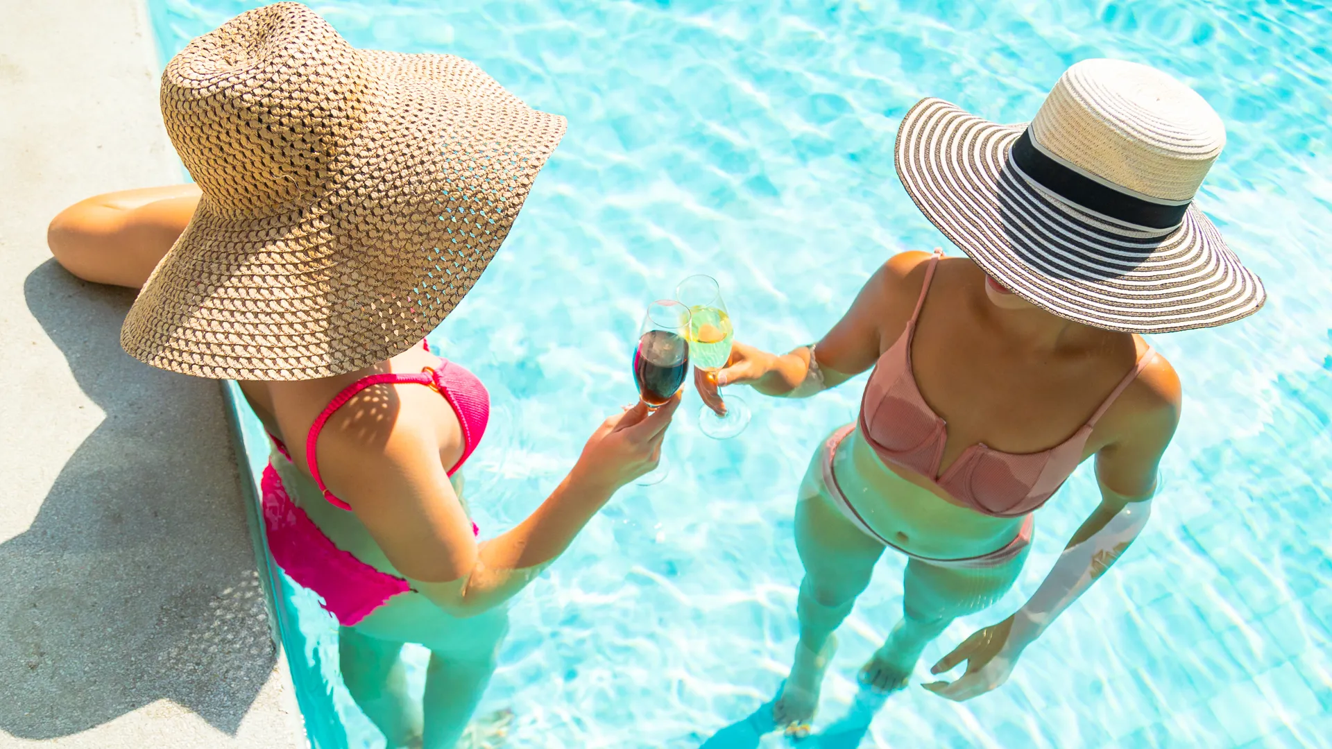 Two women in colorful bikinis toasting with wine glasses at a turquoise poolside, one sitting on the edge wearing a wide-brimmed straw hat, the other standing in the water with a striped hat, under a sunny sky.