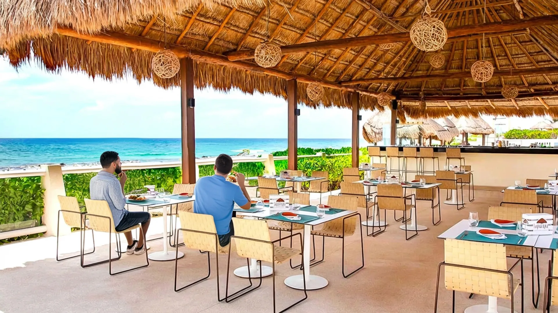Open-air beachfront restaurant at Crown Paradise Club Cancun under a thatched palapa roof with woven hanging lanterns, wooden tables set with blue placemats, plates of food, and glasses, lounge chairs nearby, lush greenery, and the turquoise Caribbean Sea visible through wooden railings under a partly cloudy sky.