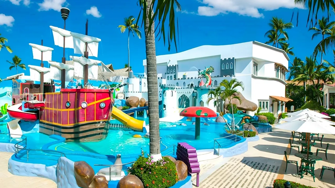 Vibrant pirate-themed children's water play area at Crown Paradise Club Cancun, including a large red ship with sails and cannons, yellow spiral slides, blue splash pools, castle-like white structures with turrets, palm trees, umbrellas, lounge chairs, and ocean views under a clear blue sky.