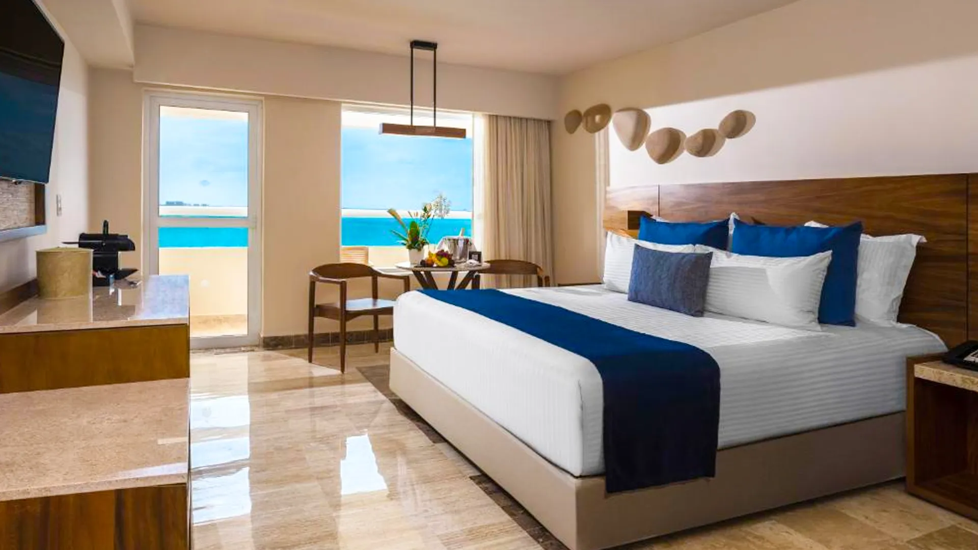 A cozy ocean-view bedroom in a Cancun resort suite, showcasing a king-size bed with white linens, blue throw pillows and runner, wooden headboard, a small table with fruit and champagne in an ice bucket, two armchairs, potted tropical plants, and sliding glass doors opening to a balcony with sea views under a clear blue sky.