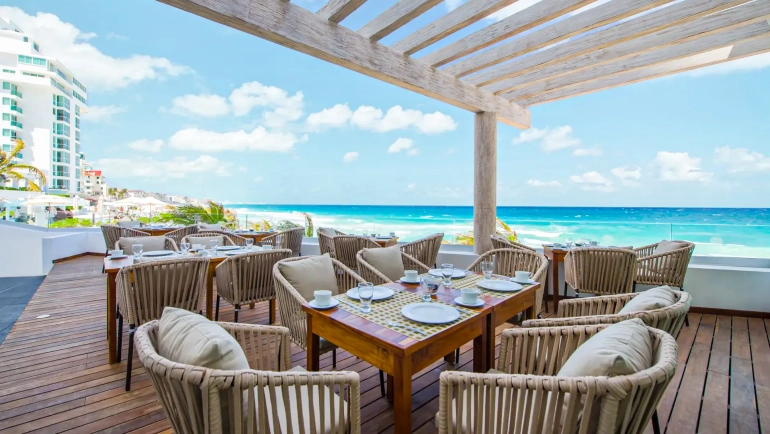 Outdoor dining terrace at Oleo Cancun Playa with ocean views, wooden pergola, and set tables overlooking turquoise waters.