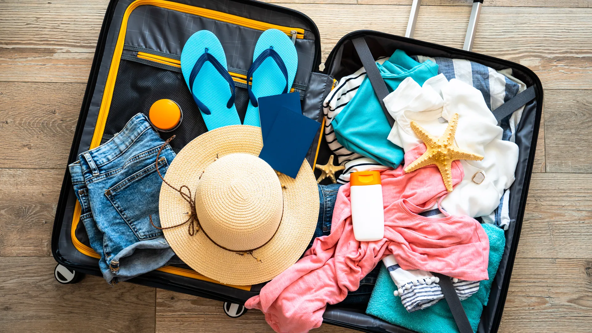 An open suitcase on a wooden floor packed with summer clothing, including a straw hat, blue flip-flops, a pink shirt, jeans, a sunscreen bottle, and a starfish decoration.