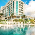 A panoramic view of the pool area at Sandos Cancun Resort featuring a large lagoon-style pool with white cabanas and red cushions, thatched-roof structures, palm trees, lounge chairs, and the turquoise ocean beyond under a vibrant blue sky.