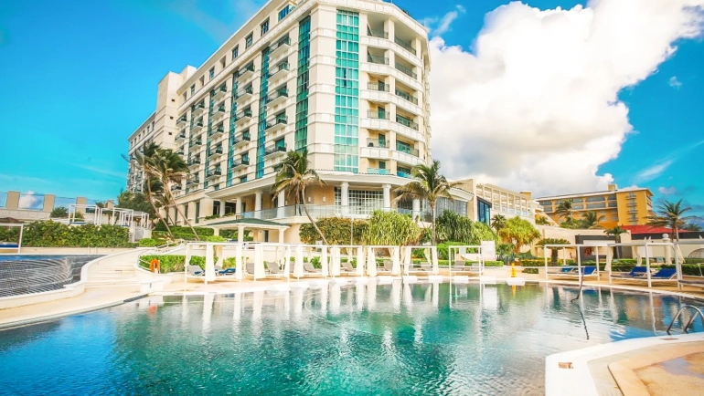 A panoramic view of the pool area at Sandos Cancun Resort featuring a large lagoon-style pool with white cabanas and red cushions, thatched-roof structures, palm trees, lounge chairs, and the turquoise ocean beyond under a vibrant blue sky.