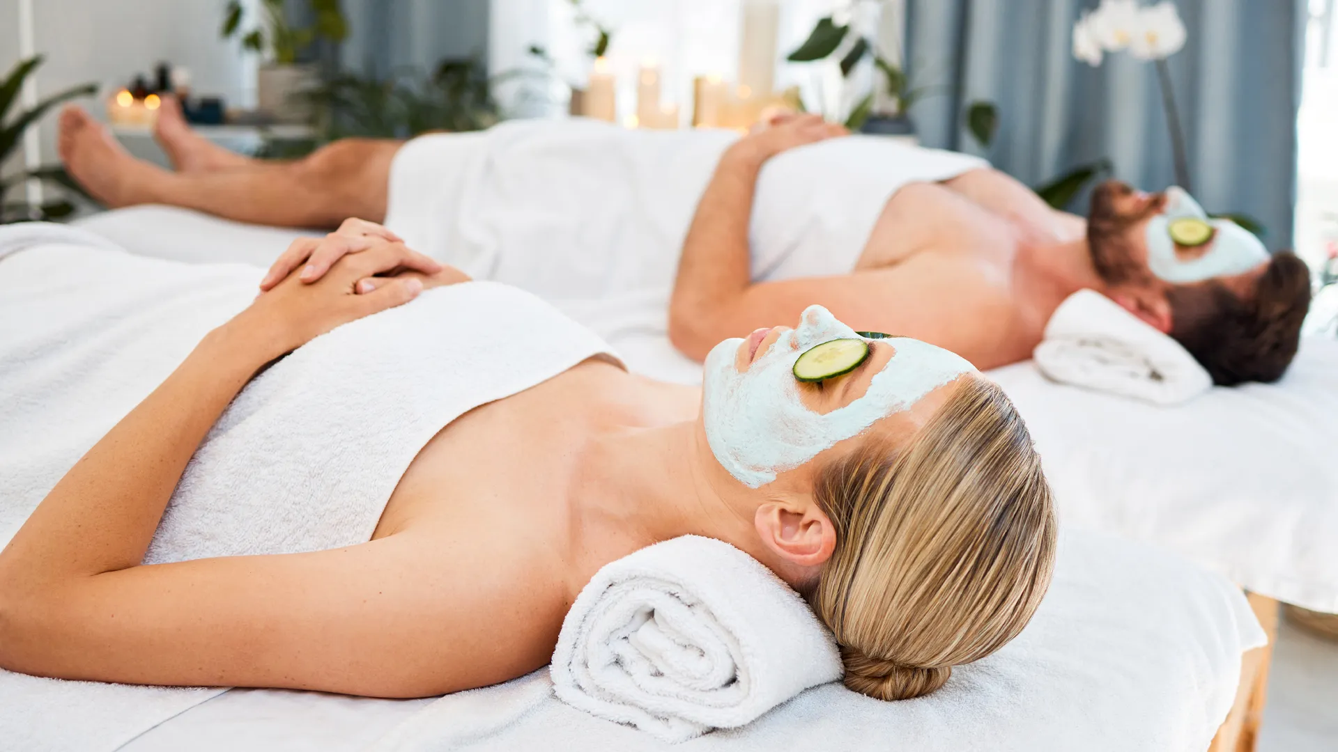 A couple lying on spa beds with green facial masks and cucumber slices on their eyes, surrounded by white towels and soft lighting with plants in the background.