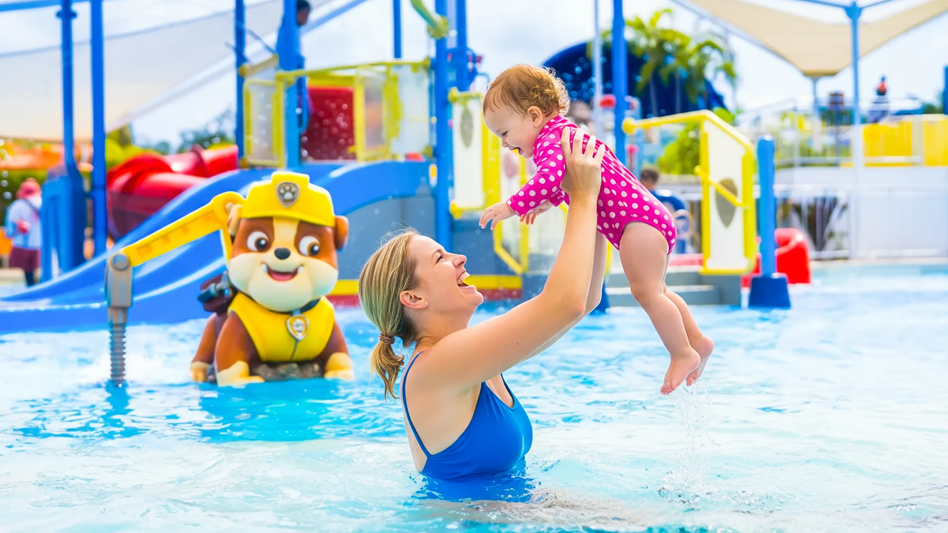 A woman in a blue swimsuit holding a laughing child in a pink polka-dot swimsuit above the water in a colorful kids' pool area with slides and a Paw Patrol character statue.