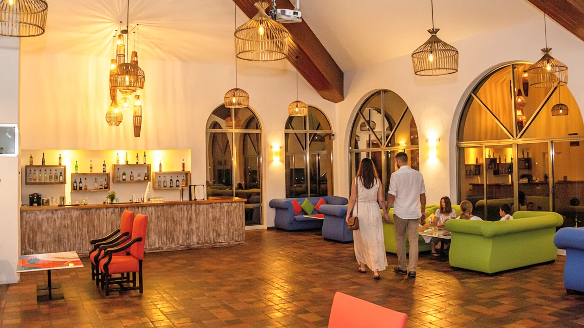 An elegant indoor resort lounge with arched windows, warm lighting from hanging lanterns, a wooden bar with bottles, colorful sofas, and people enjoying the spacious, cozy atmosphere with tiled flooring.