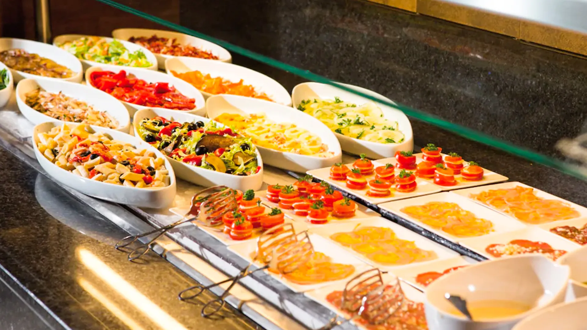 A buffet display with various dishes including pasta salads, vegetable salads, sliced vegetables, and appetizers with tomato toppings, arranged in white oval trays under a glass cover on a sleek counter.
