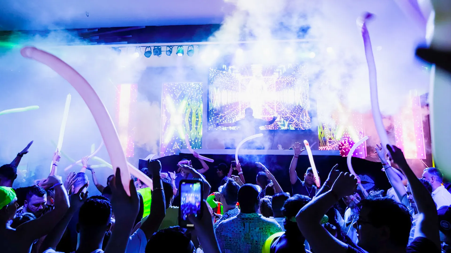 A vibrant nightclub scene with a crowd of people waving glow sticks and taking photos, a DJ performing on stage with colorful lights, smoke effects, and a large screen in the background.