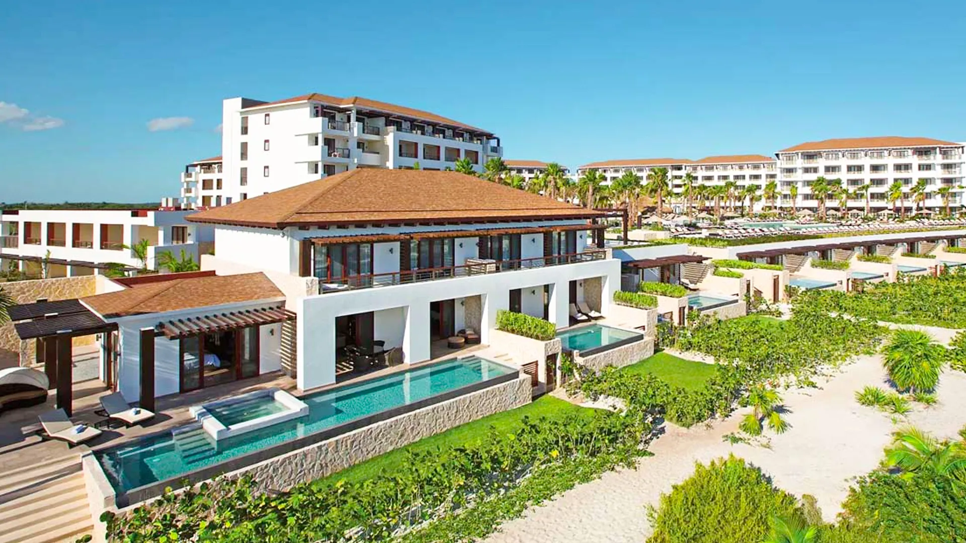 Aerial view of luxurious resort villas with terracotta roofs, private infinity pools, white buildings, palm trees, and manicured green landscapes overlooking the ocean.