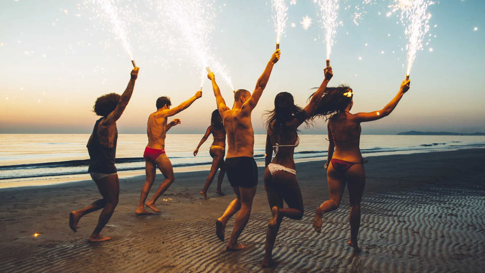 Energetic group of five young adults—three men in swim trunks and two women in bikinis—dancing on the beach at Crown Paradise Club Cancun at dusk, holding lit sparklers that trail golden sparks, silhouetted against the orange sunset sky and calm sea waves.