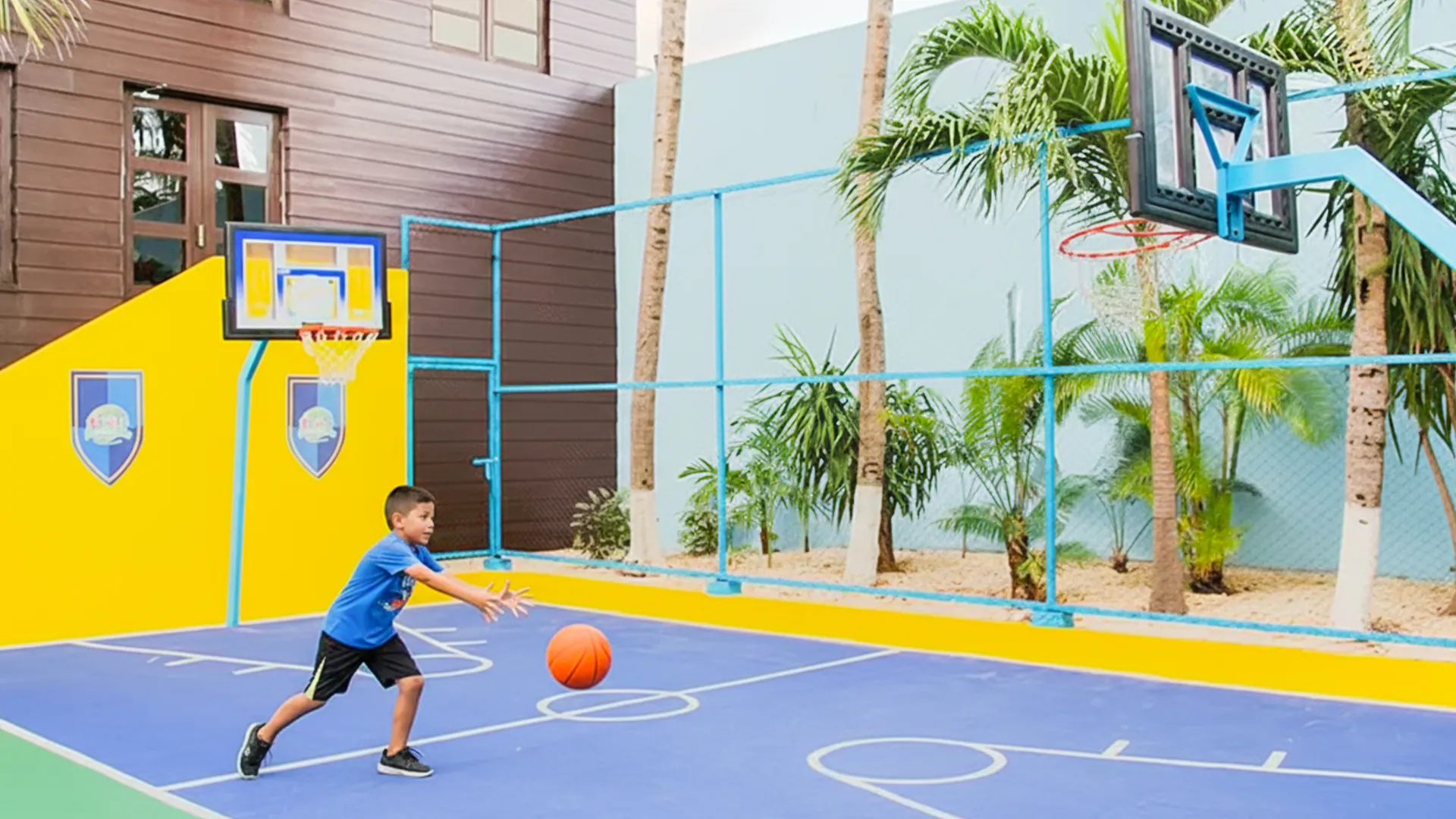 A young boy in a blue shirt and black shorts dribbling an orange basketball on an outdoor court at Crown Paradise Club Cancun, with a yellow backboard and hoop, green palm trees, and a fenced area under a clear sky.