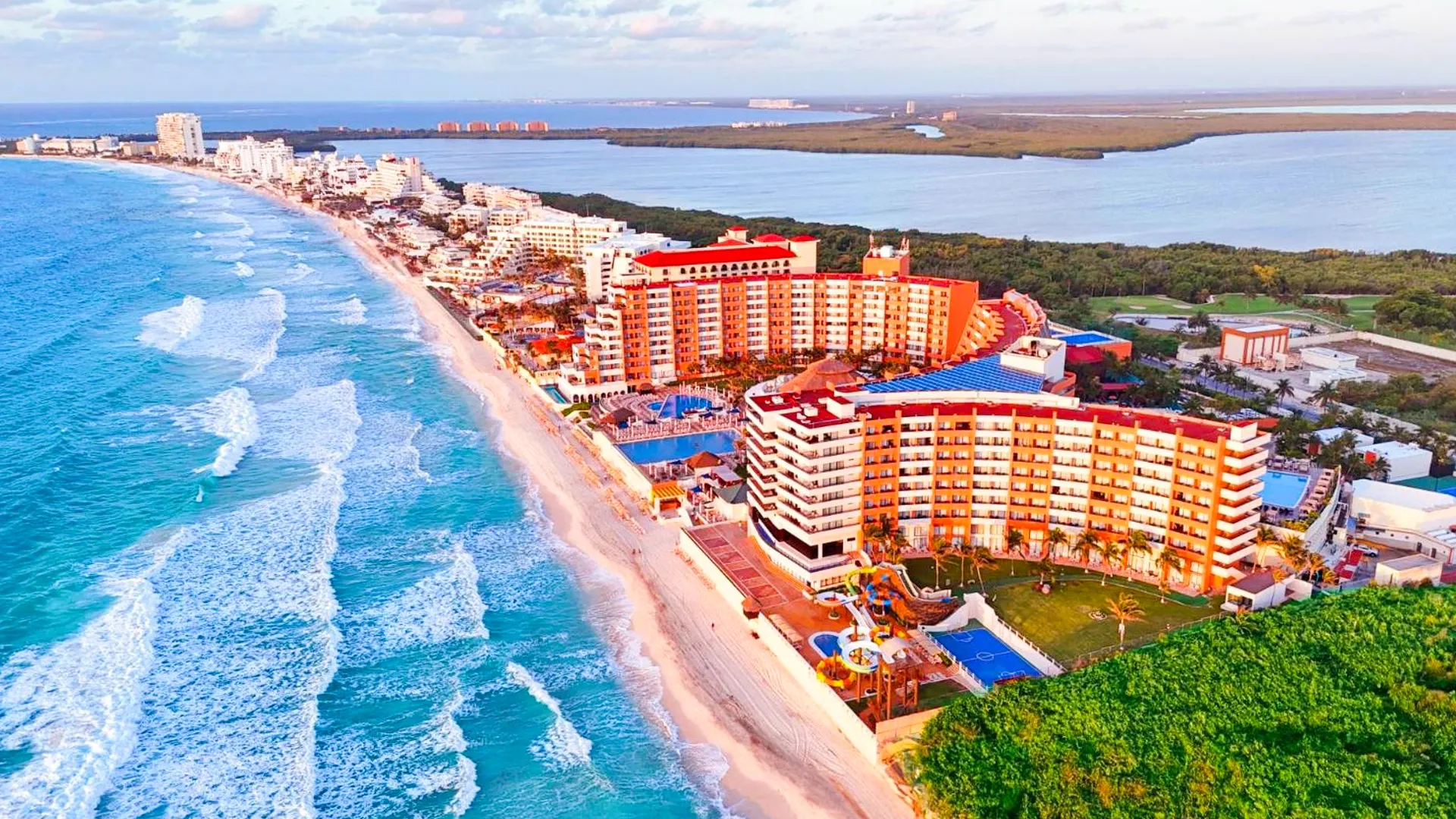 Aerial view of Crown Paradise Club Cancun's coastline, showcasing a stretch of white sandy beach, turquoise ocean waves, multiple resort buildings with orange roofs, lush greenery, and a clear blue sky.
