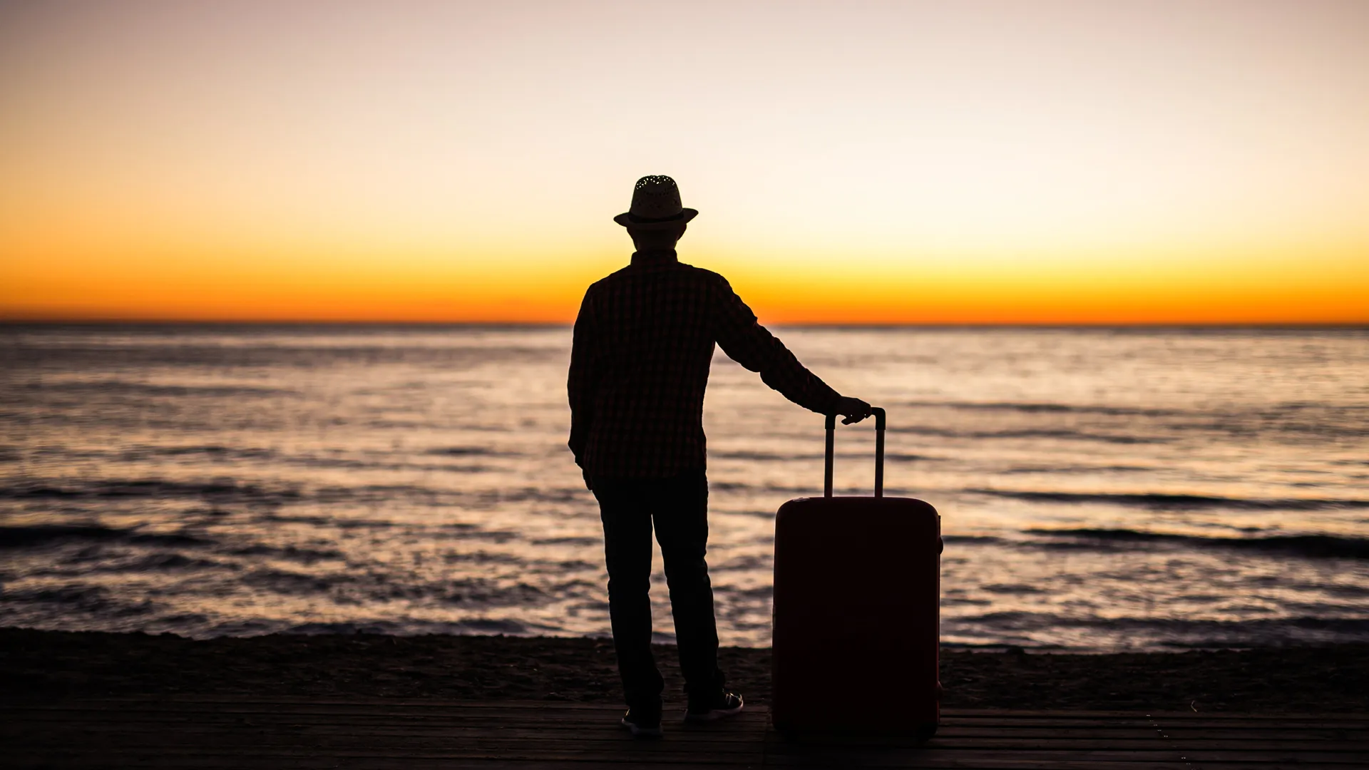 Traveler in a hat standing on the beach with a suitcase, watching the colorful sunset over the sea.