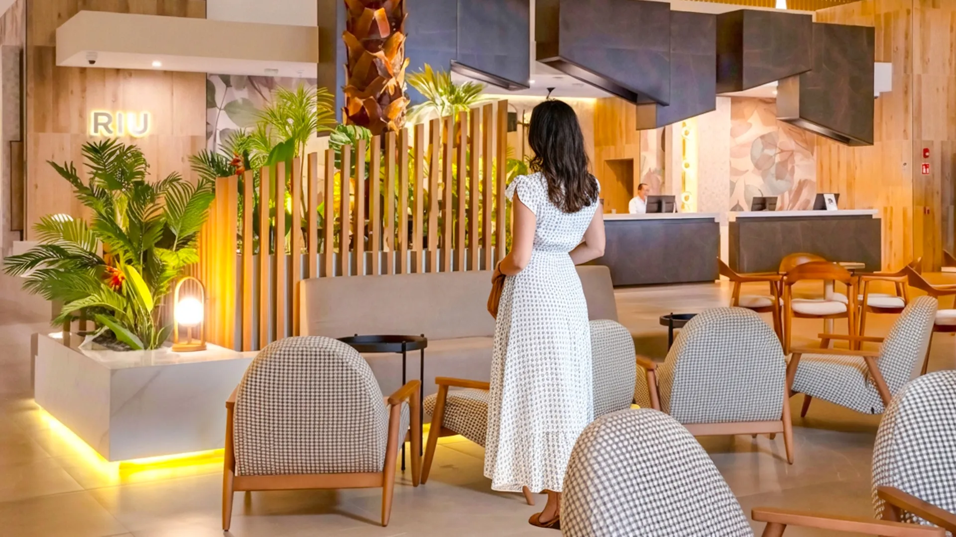  Guest walking through the stylish lobby of Riu Caribe Cancun with tropical plants and warm lighting.
