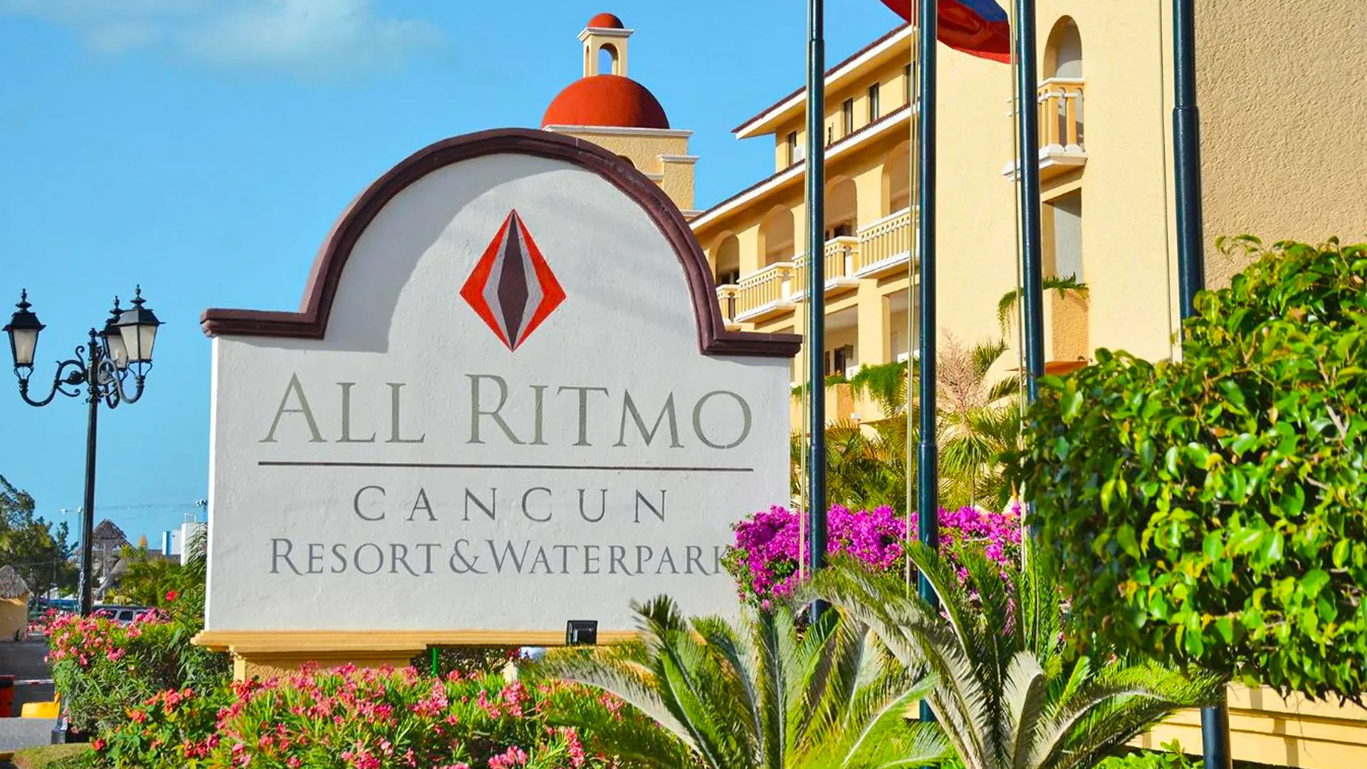Entrance sign of All Ritmo Cancun Resort & Waterpark surrounded by tropical plants and Spanish-style buildings.